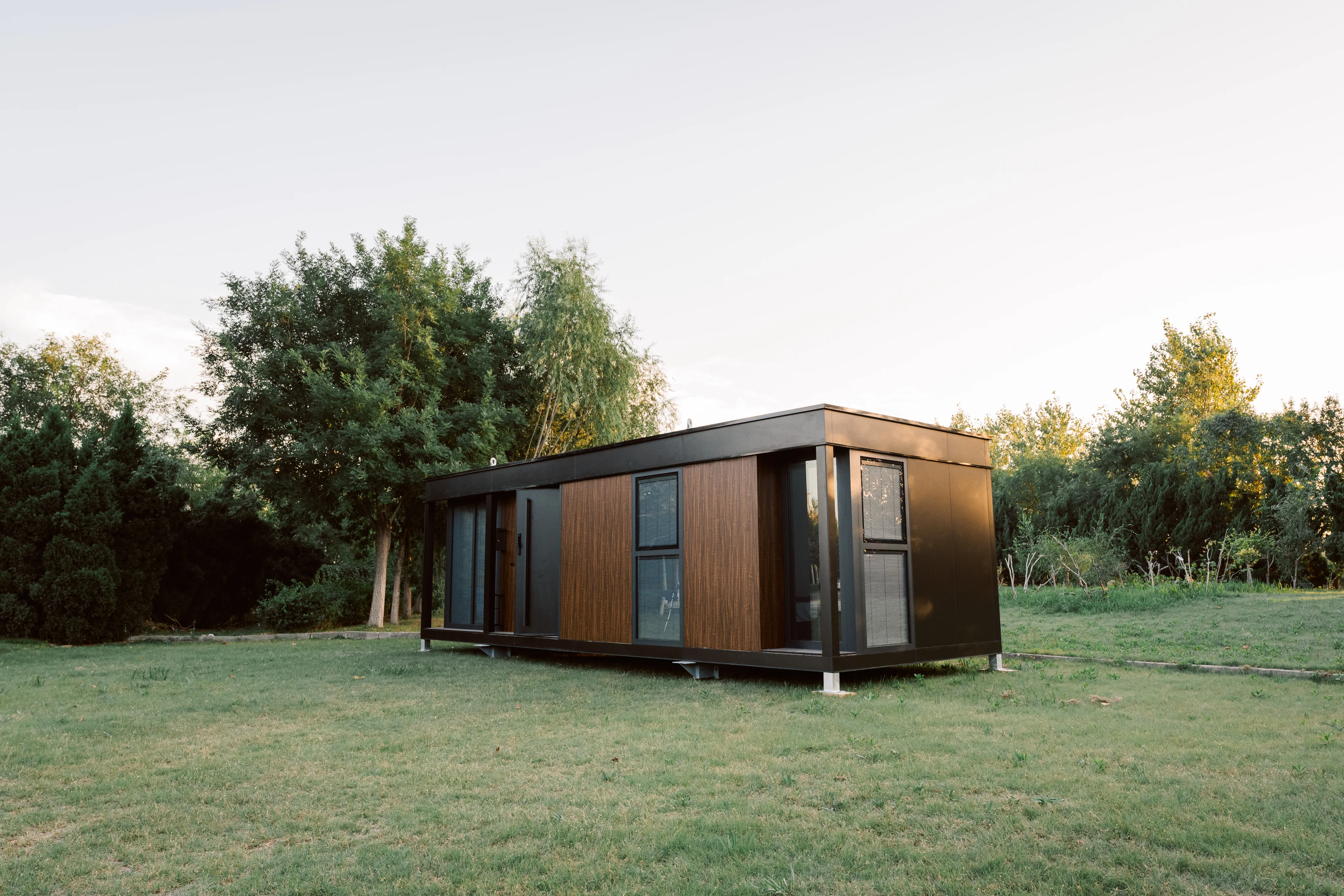Exterior view of a compact modular home with wood siding, dark trim, and surrounding greenery