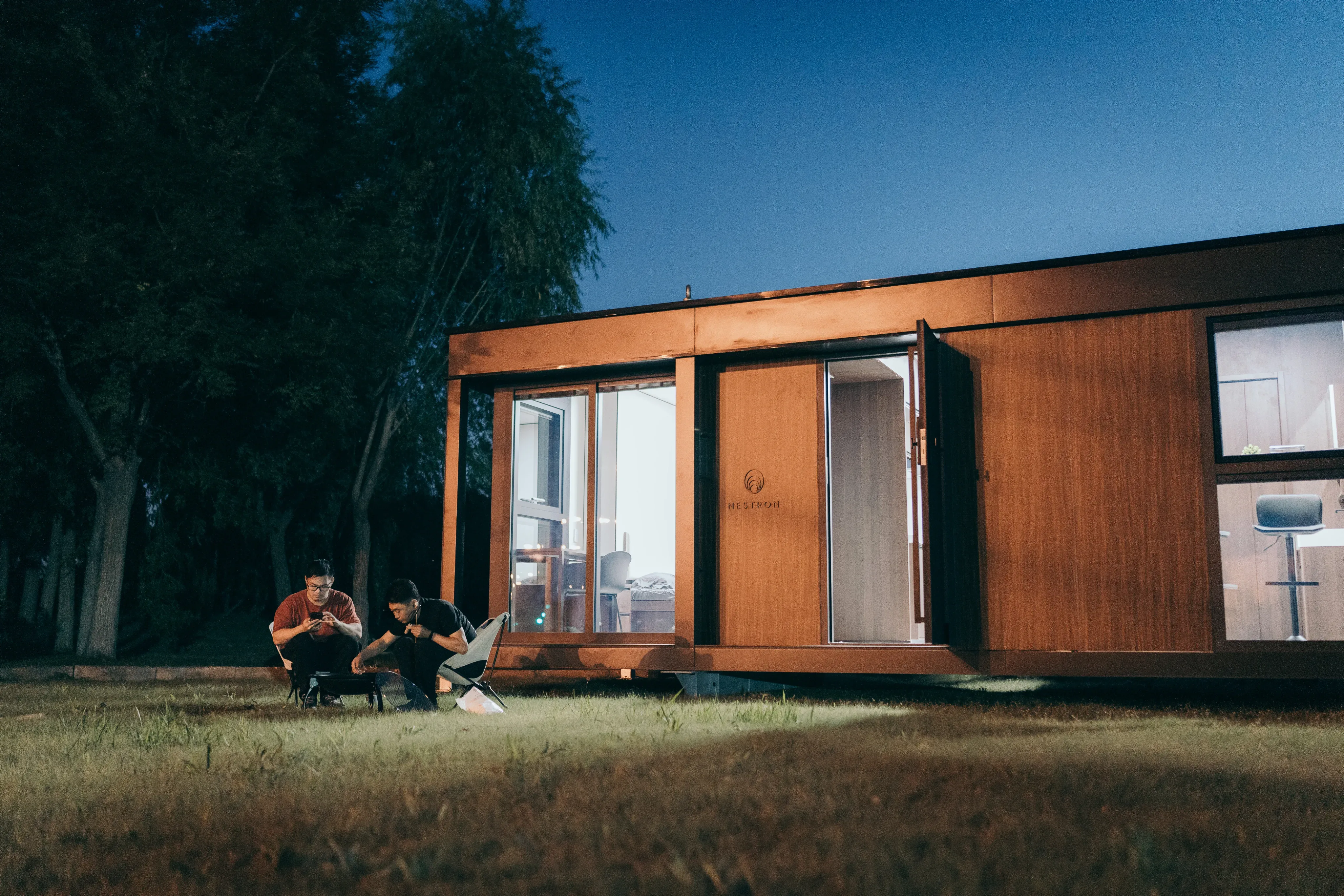 Exterior night view of a compact modular home with wood siding and warm interior lighting