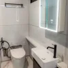 Nestron Legend One bathroom rendering with a white vanity, toilet, illuminated mirror, and tiled walls