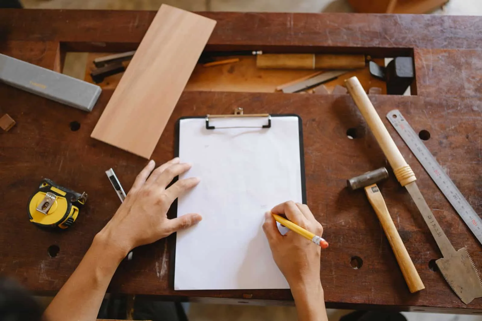 Top-down view of hands sketching plans on a clipboard surrounded by woodworking tools, measuring equipment, and wood pieces