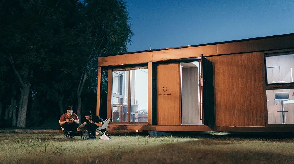 Exterior night view of a modern modular home with wood siding, large glass doors, and warm interior lighting