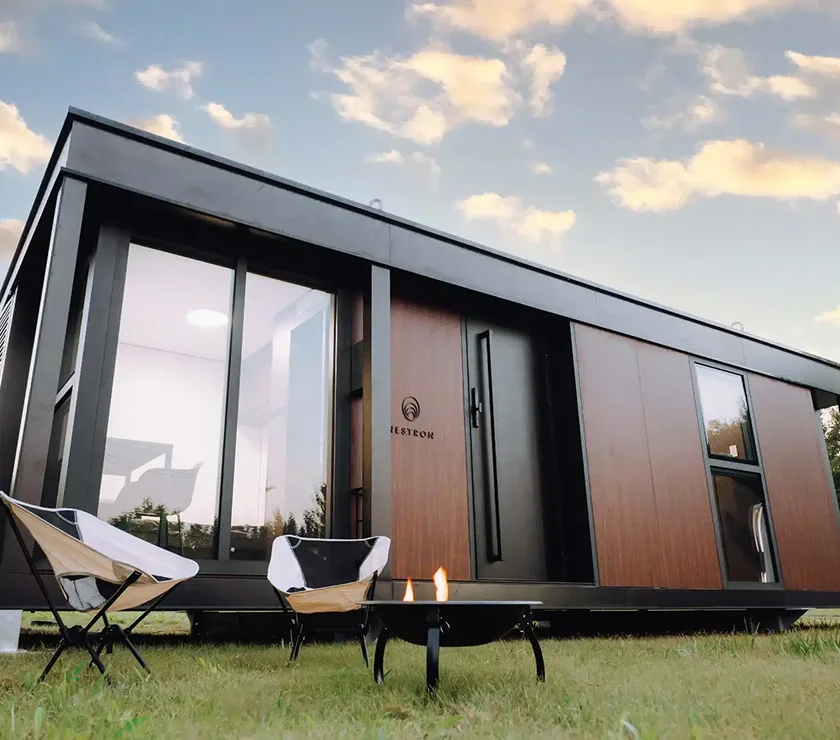 Angled exterior view of a fully assembled Nestron modular home with large windows, outdoor chairs, and fire pit